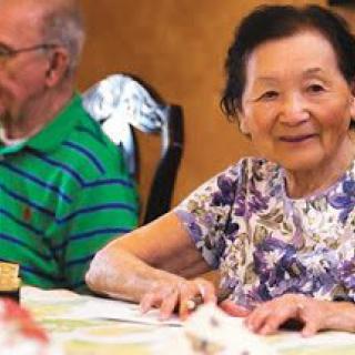 An elderly couple sitting at a dining table, with the woman smiling and wearing floral attire.