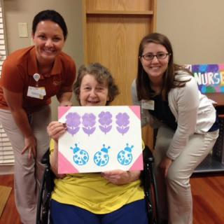 A smiling woman in a wheelchair proudly displays her artwork, accompanied by two women in a healthcare setting.