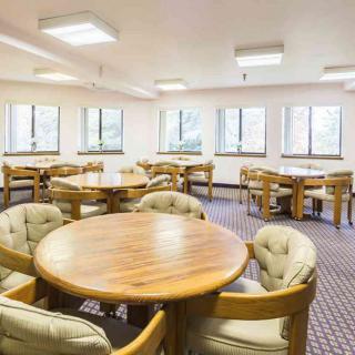 A spacious dining area with multiple tables and chairs, featuring large windows and carpeted flooring.