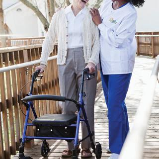 A caregiver assists an elderly person using a walker on a wooden ramp.