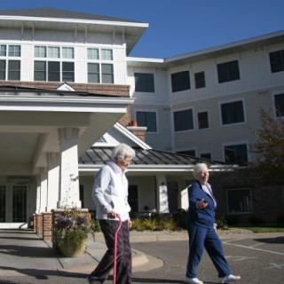 Two elderly individuals walking outside a retirement home on a sunny day.