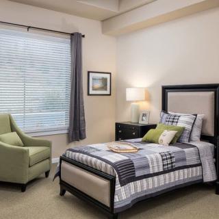 A cozy bedroom featuring a single bed with a patterned quilt, a green armchair, and soft lighting from a table lamp.