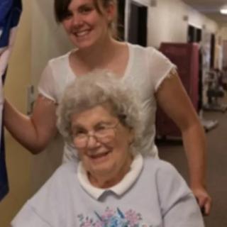 A smiling elderly woman sitting in a wheelchair with a younger woman standing behind her indoors.
