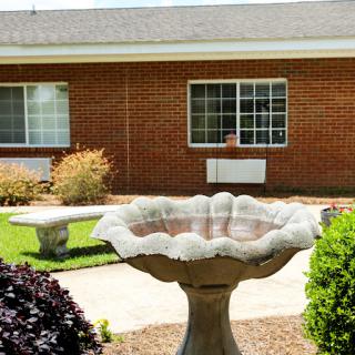A serene garden area featuring a decorative birdbath and lush greenery surrounding a brick building.