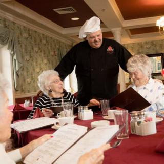 A chef interacts with elderly guests at a restaurant table, discussing the menu.