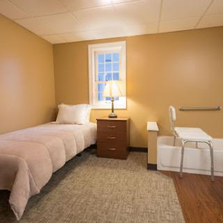 A cozy bedroom featuring a single bed, a nightstand with a lamp, and a chair, all set against warm-colored walls.