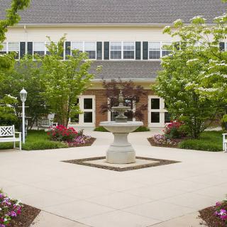 A peaceful courtyard with a central fountain, surrounded by benches, greenery, and flowers in front of a building.