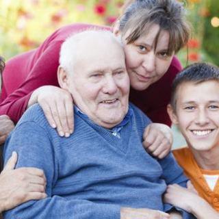 A family gathering with an elderly man surrounded by loved ones, smiling outdoors.