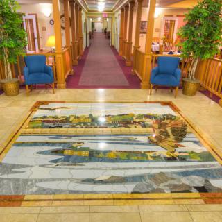 A hotel lobby featuring a decorative floor artwork, flanked by potted plants and blue chairs along the hallway.