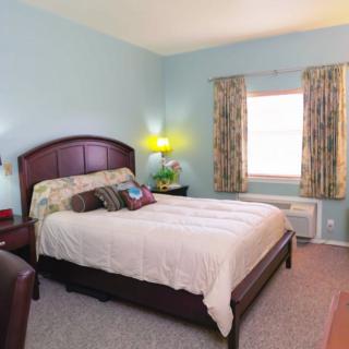 A cozy bedroom with a large bed, wooden headboard, and soft pillows. A nightstand with a lamp is beside the bed, and curtains cover the window, creating a warm atmosphere.
