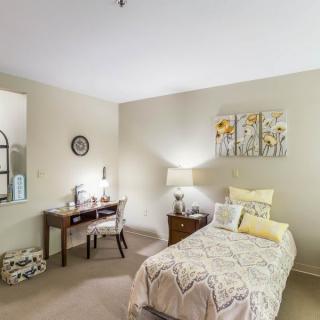 A cozy bedroom featuring a single bed, a desk with a chair, decorative artwork, and a bedside lamp.