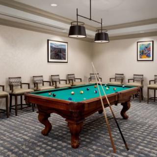 A well-lit game room featuring a billiard table surrounded by chairs, with cue sticks resting against it.