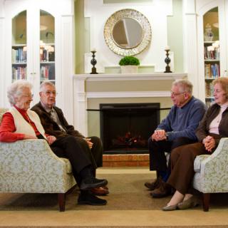A group of seniors sitting in a living room having a conversation near the fireplace.