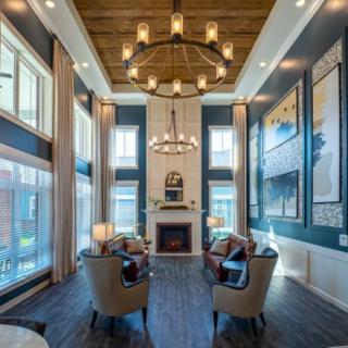 A luxurious and spacious living room featuring high ceilings, large windows, elegant chandeliers, and a central fireplace surrounded by comfortable seating.