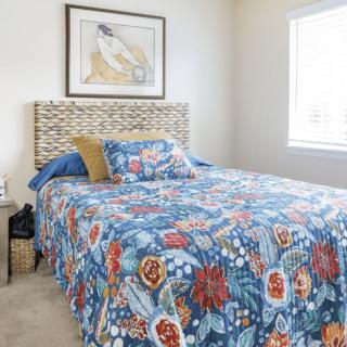 A cozy bedroom with a bed adorned in colorful floral-patterned bedding, complemented by framed artwork and natural light streaming through the window.