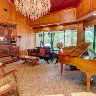 A luxurious living room with a grand piano and elegant chandelier, featuring wood paneling and large windows.