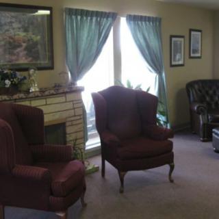 A cozy living room with two upholstered chairs, a fireplace, green curtains, and framed art on the walls.