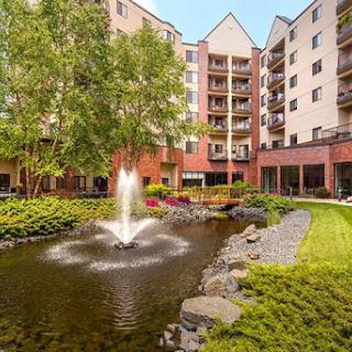 A serene view of a landscaped pond featuring a water fountain, surrounded by an apartment complex.