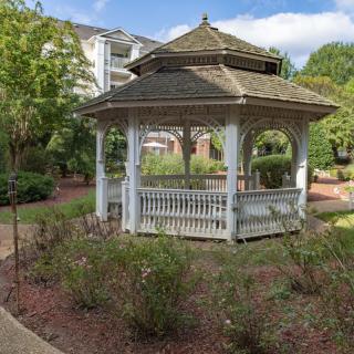 A charming wooden gazebo surrounded by a well-maintained garden area, set against a backdrop of trees and residential buildings.