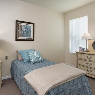 A cozy bedroom with a single bed, featuring decorative pillows and a neatly arranged dresser with a lamp.