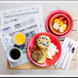 A breakfast spread with a newspaper, featuring sausage, potatoes, eggs, a muffin, fruit salad, coffee, and orange juice.