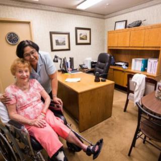 A caregiver providing support to an elderly person in a wheelchair within an office setting.