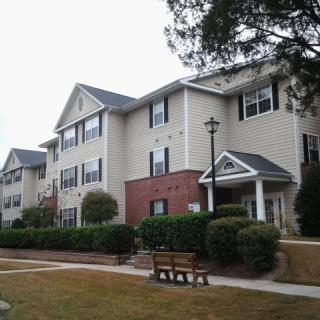 A suburban apartment building with surrounding greenery and a bench in the foreground.