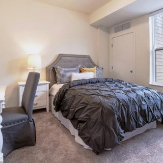 A cozy bedroom featuring a neatly made bed with dark bedding, a desk with a chair, and a lamp providing light.