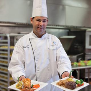 A chef presents two elegantly plated dishes in a professional kitchen.