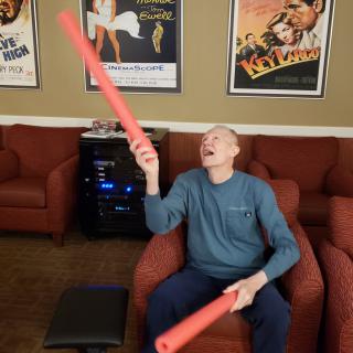 A person sitting on a chair indoors, playfully holding two foam noodles with movie posters in the background.