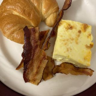 A breakfast plate featuring crispy bacon, a croissant, and a square of egg casserole.