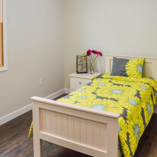 A small bedroom featuring a single bed with bright yellow bedding, complemented by a decorative pillow and a side table with a lamp and flower vase.