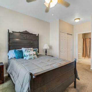 A cozy bedroom featuring a wooden bed with decorative pillows, a bedside lamp, and a ceiling fan. An open door leads to an adjacent room.