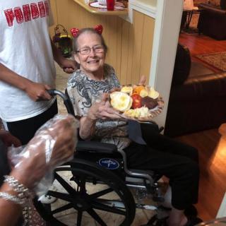 An elderly woman in a wheelchair is holding a birthday cake and smiling during a celebration.