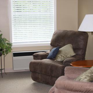 A cozy living room with a comfortable armchair and sofa, adorned with cushions, next to a window and a lamp.