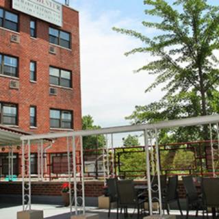 An outdoor patio area with tables and chairs next to a brick building, surrounded by trees under a clear sky.