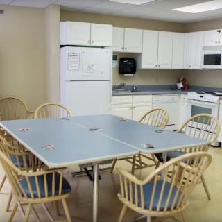 A communal kitchen area with a large dining table and wooden chairs, surrounded by white cabinets and appliances.