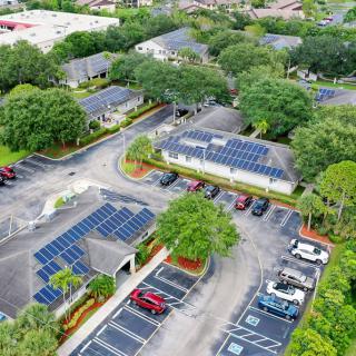Aerial view of a complex with solar panels installed on rooftops and parking lot canopies, surrounded by greenery.