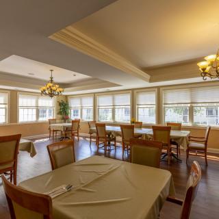 A well-lit dining area with neatly arranged tables and chairs, featuring large windows and elegant chandeliers.