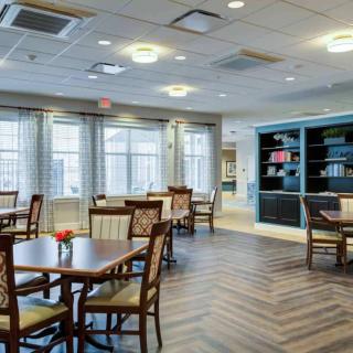 A modern dining area with tables and chairs, complemented by a bookshelf and large windows.