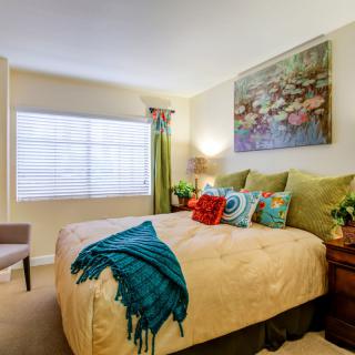 A cozy bedroom with colorful decor, featuring a large bed adorned with a variety of pillows and a vibrant blue throw blanket. The walls are decorated with a large piece of artwork above the headboard, and natural light streams in through a window.