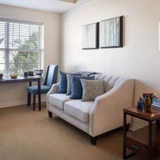 A cozy living room with a white sofa, blue pillows, and a small dining area by the window.