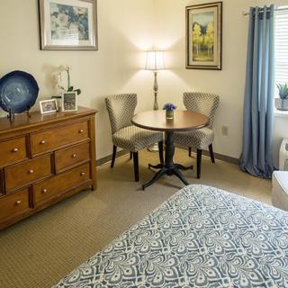 A cozy bedroom with a dresser, round table, and two chairs, adorned with decorative artwork and a floor lamp.
