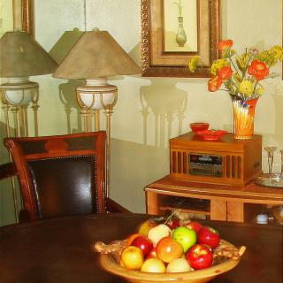 A cozy living room setup featuring a table with a fruit bowl, decorative flowers, lamps, and artwork on the wall.