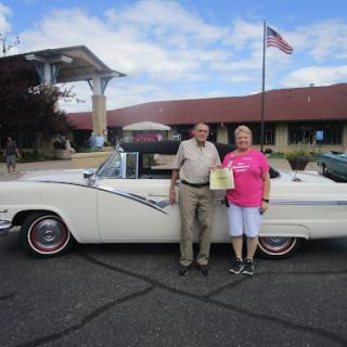 A couple celebrates their classic car at an outdoor event, proudly displaying a certificate next to their vintage vehicle.