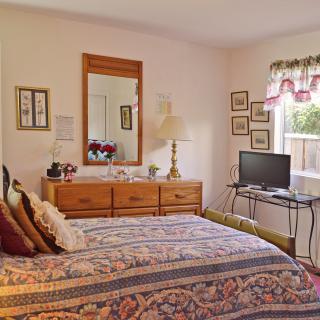 A cozy bedroom with a bed, dresser, and TV, featuring floral decor and natural light.