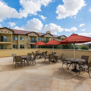 An outdoor patio with tables, chairs, and red umbrellas set against a building exterior under a partly cloudy sky.