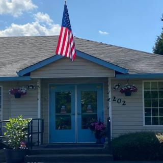 A small house with blue trim and an American flag flying above the front door, surrounded by bushes and flowers on a sunny day.