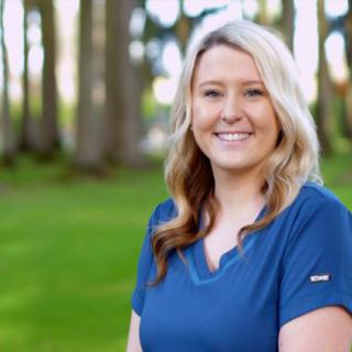 A woman with blonde hair smiling outdoors in a natural setting with trees in the background.