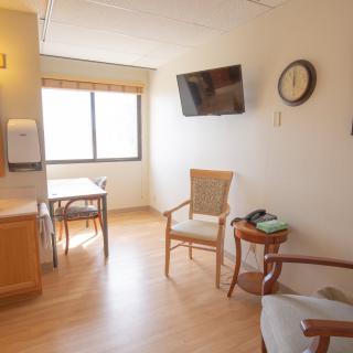 A well-lit room with a table, chairs, wall-mounted television, and a clock.